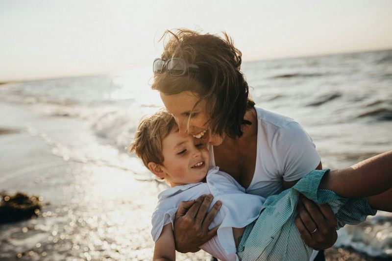 A mother laughing as she lifts her young son on the beach